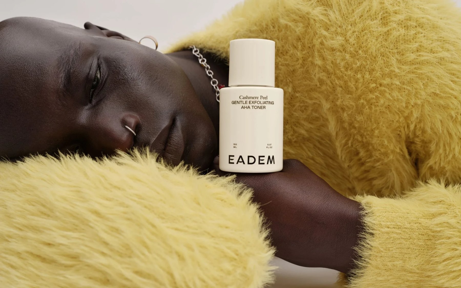 Woman resting head while displaying Eadem skincare bottle.