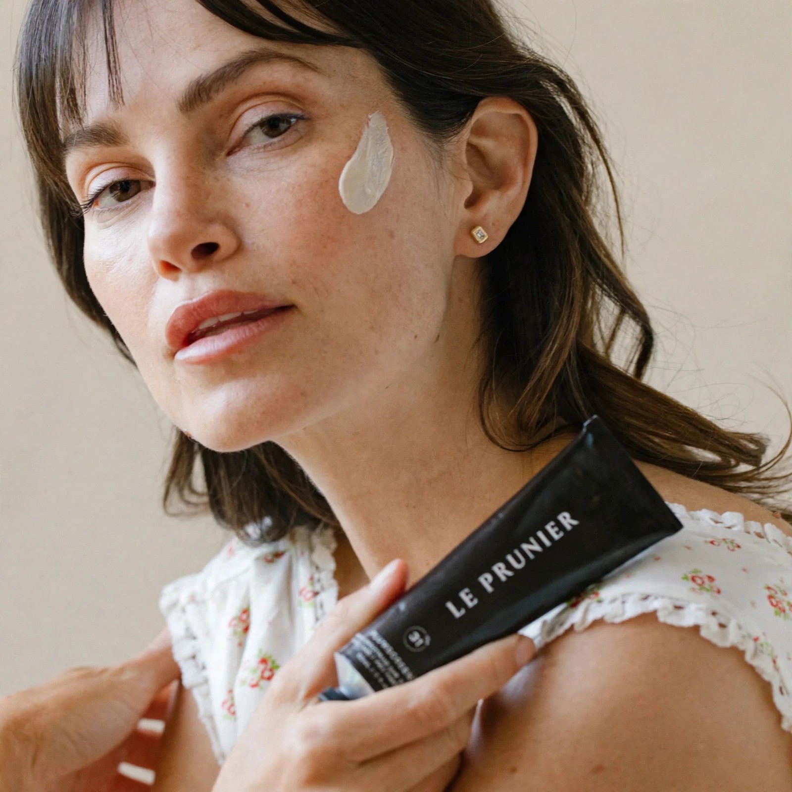 Model displays Plumscreen tube while wearing sunscreen on cheek.
